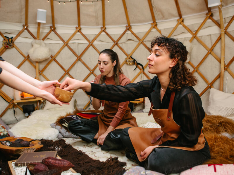 Two women are sitting in a bell tent on rugs at a cacao ceremony